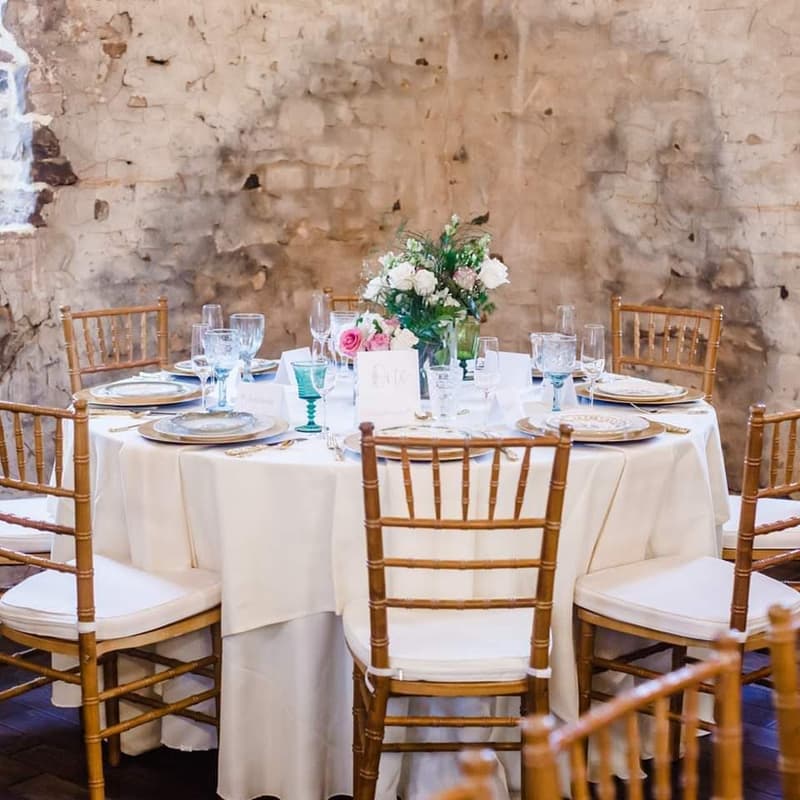 Round table with white linen and gold chiavari chairs against a stone wall