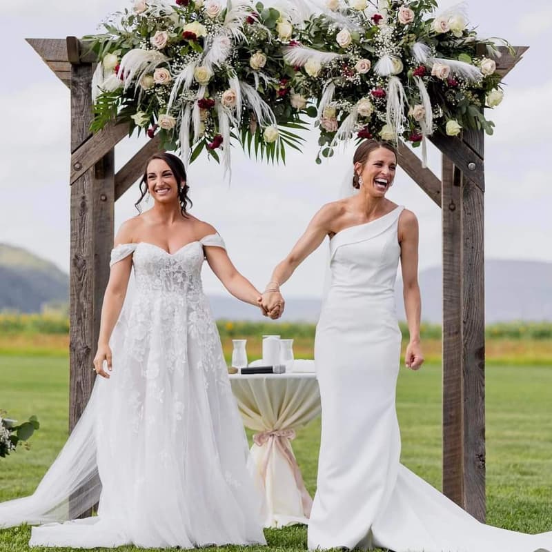 Two brides holding hands beneath a floral arch with mountains behind them